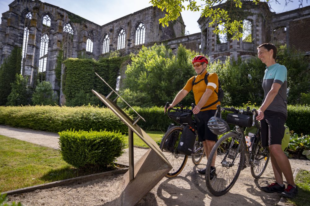 Two cyclists read an information board in front of the ivy-clad ruins of Aulne Abbey on a sunny day.