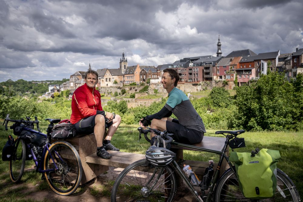 Two cyclists sit on benches with their bikes, enjoying a view of the historic town of Thuin.