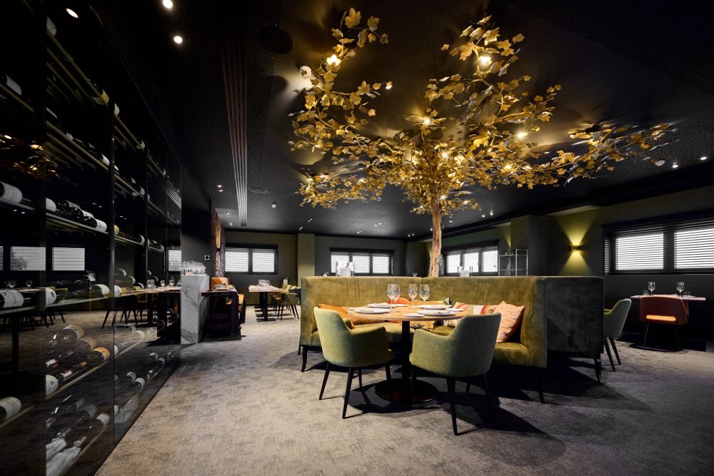 Elegant dining room with soft green seating, wine racks along the wall, and a golden tree installation with illuminated leaves as a central ceiling decoration.