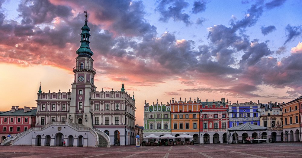 Twilight view of Zamość Market Square, featuring colorful historic buildings and a prominent clock tower amidst a dramatic sky.