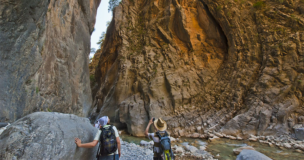 Two hikers explore a narrow canyon, capturing the towering rock walls and tranquil river below amidst lush greenery.