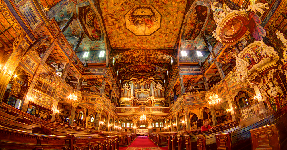 Interior of an ornate church featuring intricate paintings, wooden pews, chandeliers, and a grand organ, bathed in warm light.