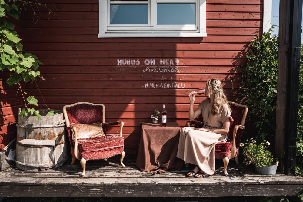 Woman enjoying a glass of rosé at Muhu Winehouse in Estonia, sitting by a rustic red wooden house.