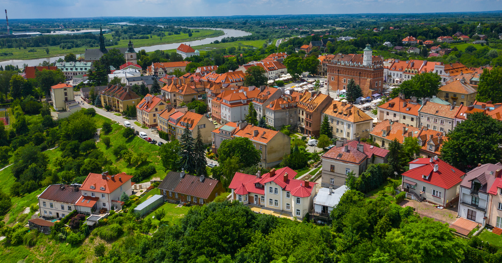 Aerial view of a picturesque town with red-roofed houses, green trees, and a river in the background under a bright blue sky.