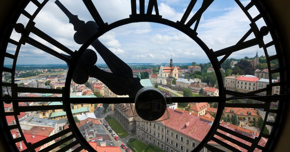 View through a large clock face, revealing a picturesque town with red-roofed buildings and lush green hills under a cloudy sky.
