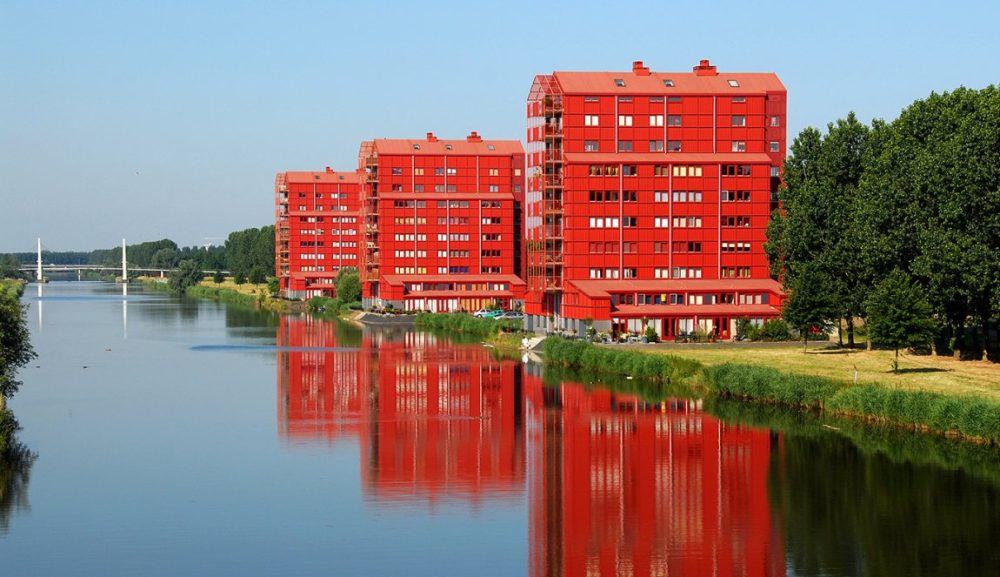 Three red modern apartment buildings reflect in a serene river, surrounded by greenery and a clear blue sky.
