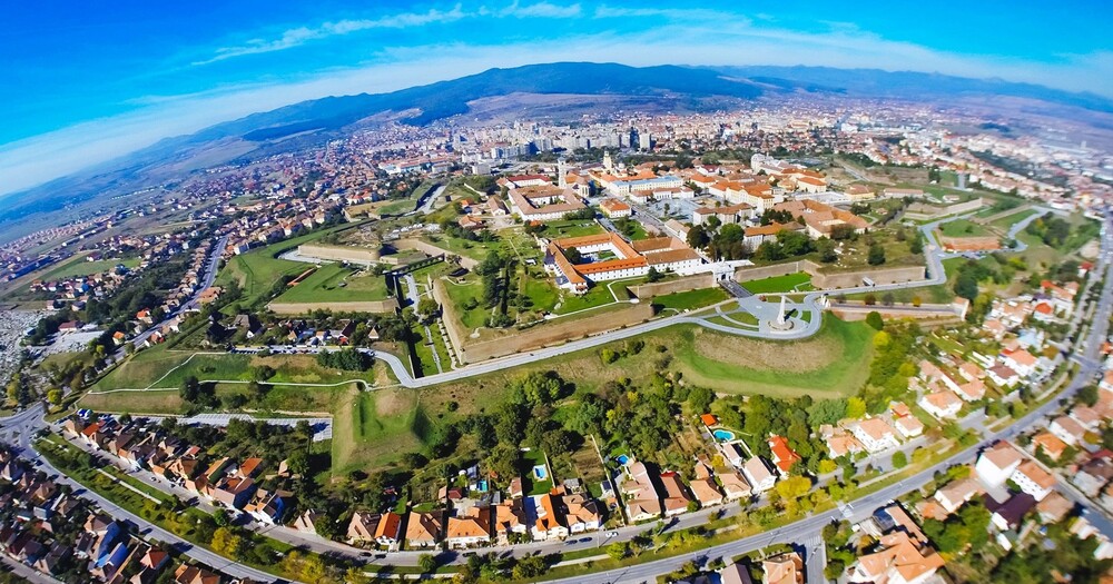 Aerial view of a city featuring green spaces, historic buildings, and residential neighborhoods against a backdrop of mountains and blue sky.