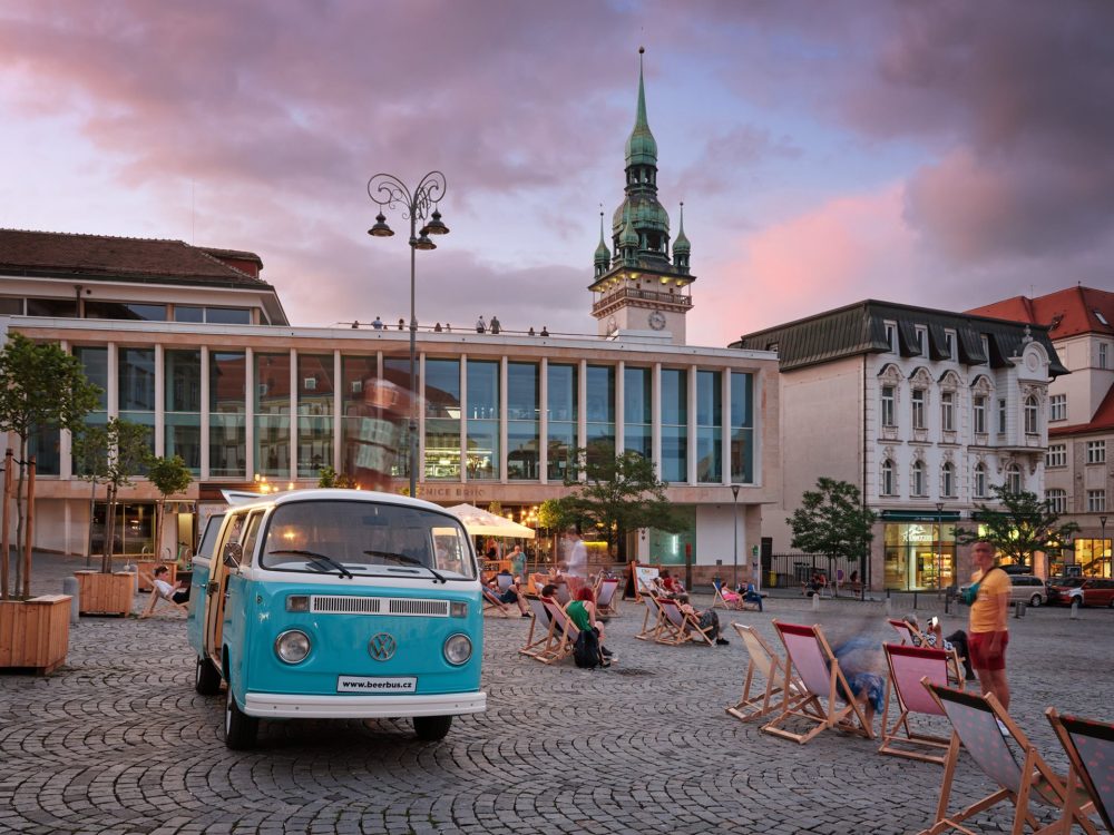 Retro blue VW van and people relaxing on deck chairs at Zelný Market in Brno during a summer evening, with the old town hall tower in the background.