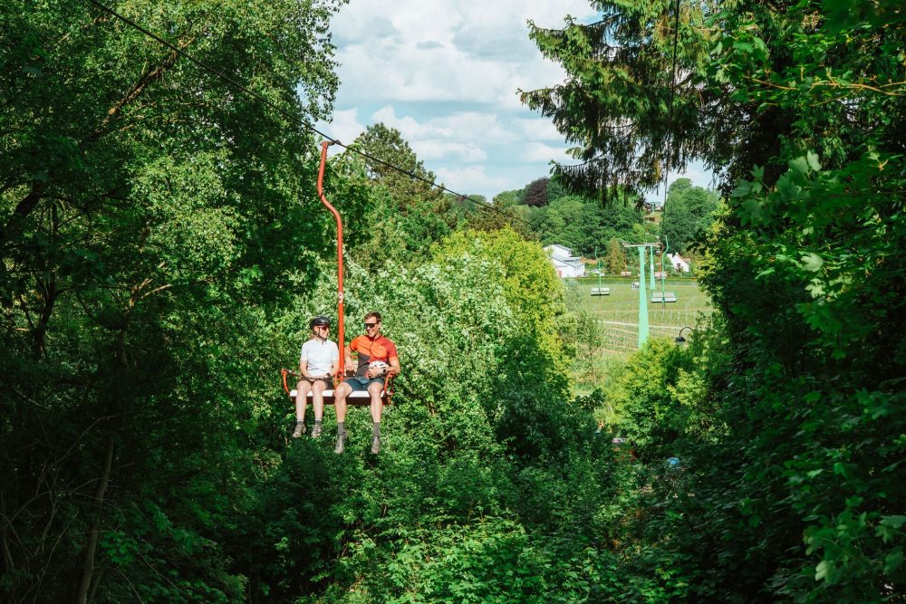 Two people on a chairlift above green vineyards and forest.