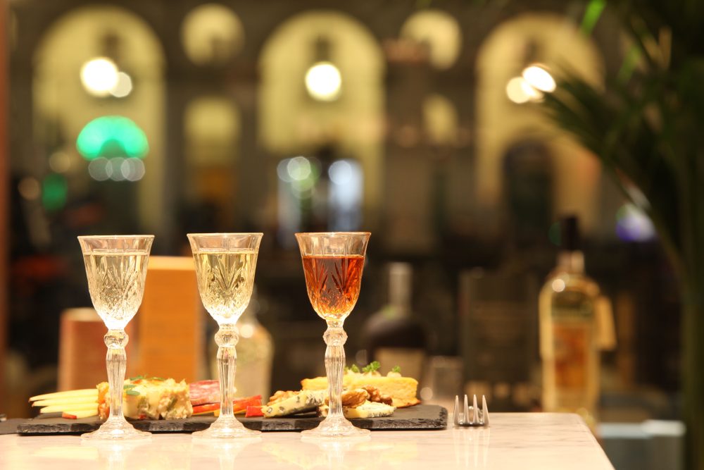 Three elegant crystal glasses filled with colorful drinks sit on a marble table, accompanied by gourmet appetizers in a softly lit venue.