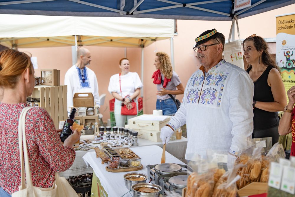 Man in traditional embroidered shirt serving customers at a farmers’ market stall with local products in Bátovce, Slovakia."