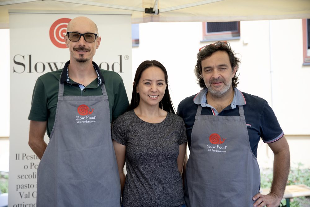 Three people smiling at a Slow Food del Pordenonese event, wearing aprons and standing in front of the organisation’s banner.