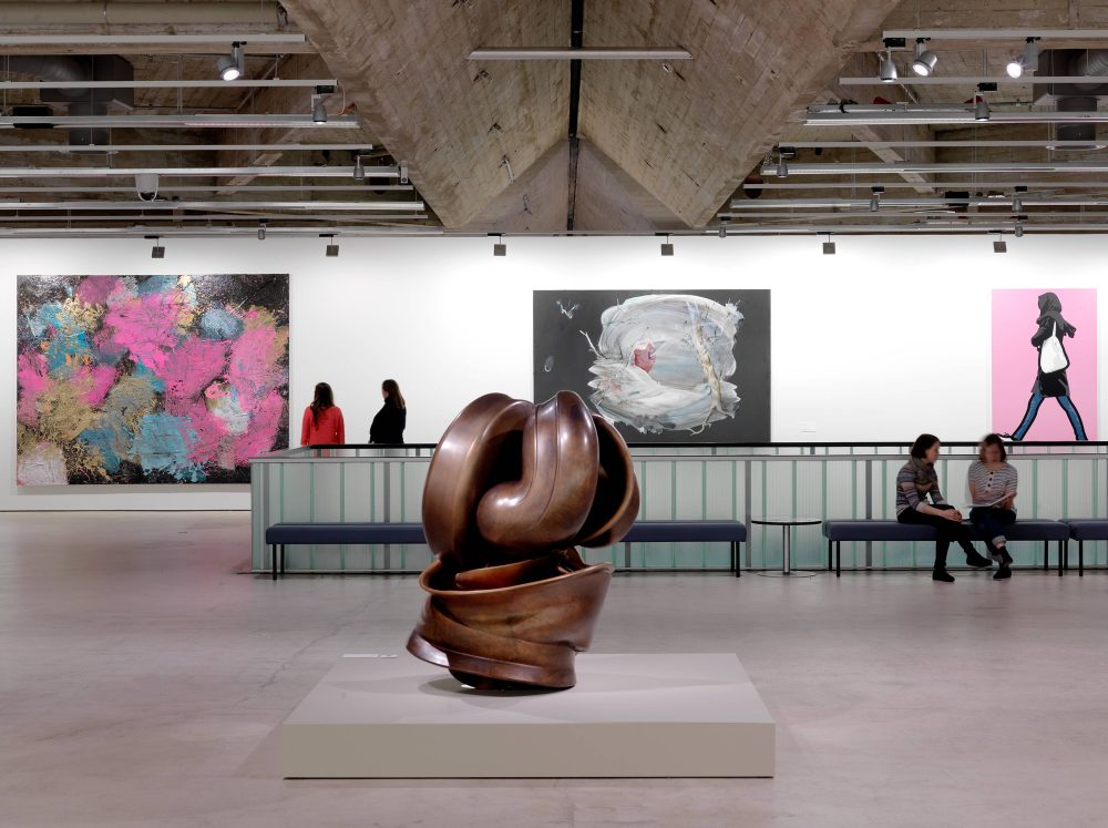 A contemporary art gallery showcasing a bronze sculpture in the foreground and colorful abstract paintings on the walls.