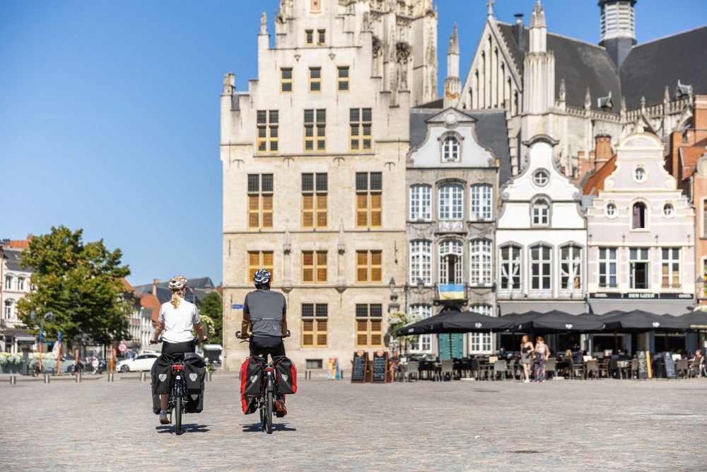 Two cyclists view historic buildings in a vibrant square on a sunny day, with cafés and greenery in the background.