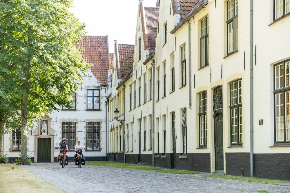 Two cyclists ride along a cobblestone path flanked by charming, historic buildings and a lush green tree in bright sunlight.