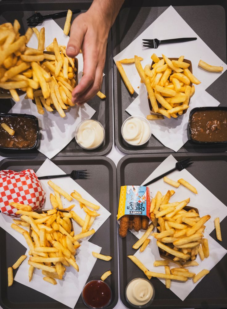 A top-down view of four trays filled with French fries, a burger, sauces, and chicken tenders, with a hand pouring more fries.