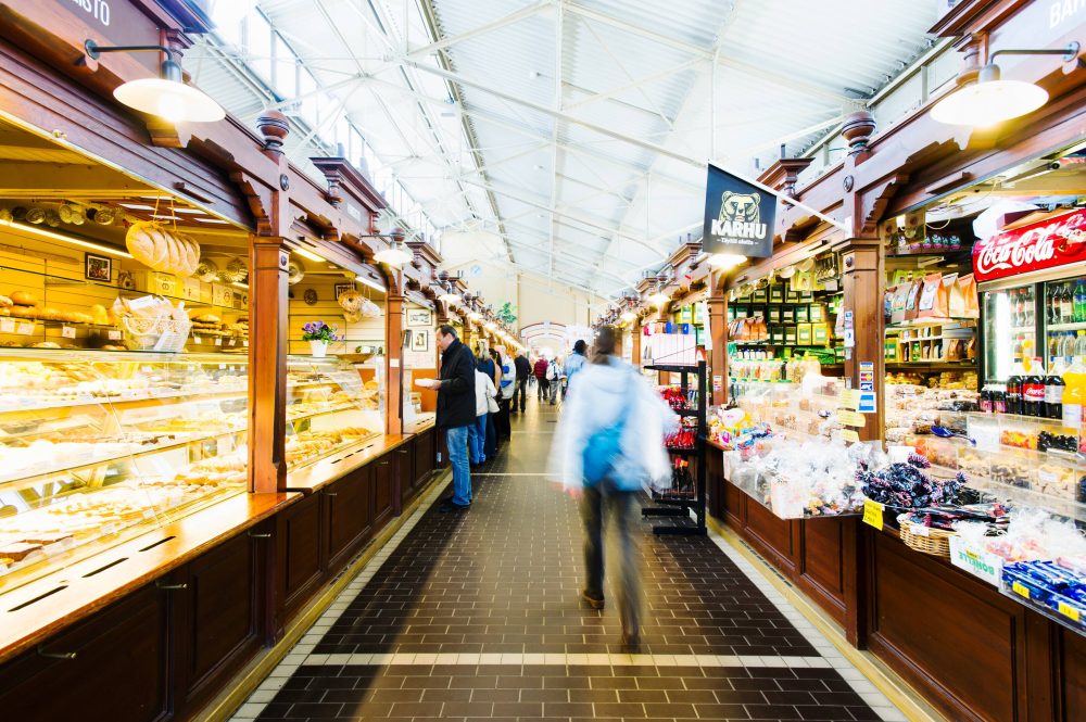 A bustling market interior with wooden stalls filled with baked goods and snacks, and shoppers exploring the vibrant offerings.