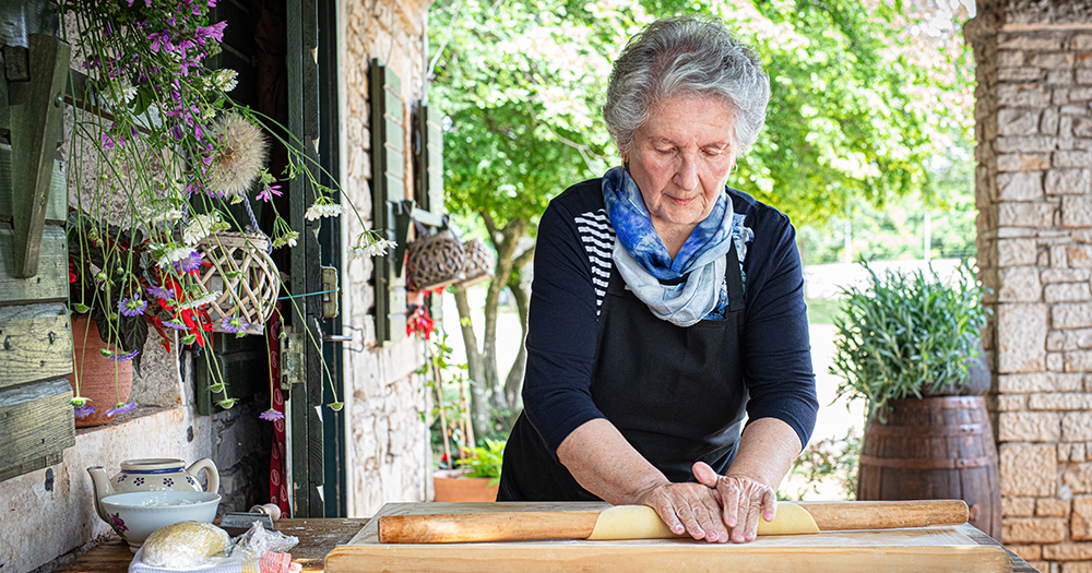 Elderly woman rolling fresh homemade pasta dough by hand in a traditional Croatian village setting.