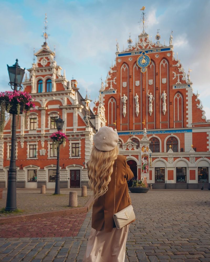 A woman with long blonde hair stands in Riga’s historic Old Town, gazing at the ornate red-and-white House of the Black Heads, a masterpiece of Gothic architecture, under a soft golden sky.