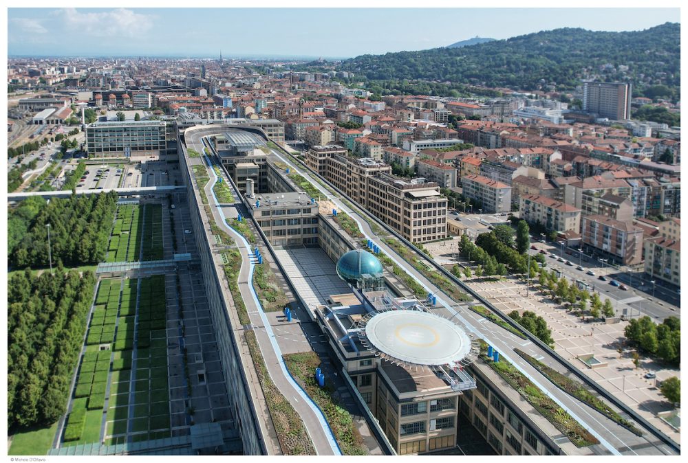 Aerial view of a modern urban landscape featuring green rooftops, buildings, and a helicopter pad, with hills in the background.