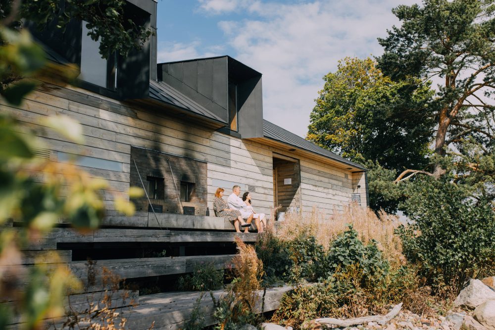 A modern wooden house with a sloped black roof, surrounded by greenery, where a group of people enjoys a sunny day on the steps.