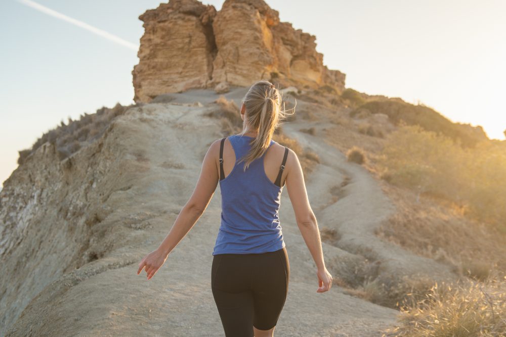 A woman walks along a narrow dirt trail through arid terrain, heading toward rocky outcrops under soft evening light.
