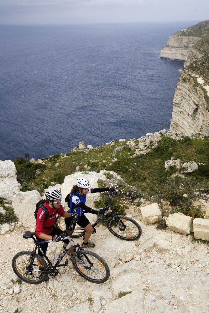 Two mountain bikers push their bikes uphill along a rocky coastal path, with steep cliffs and deep blue sea in the background.