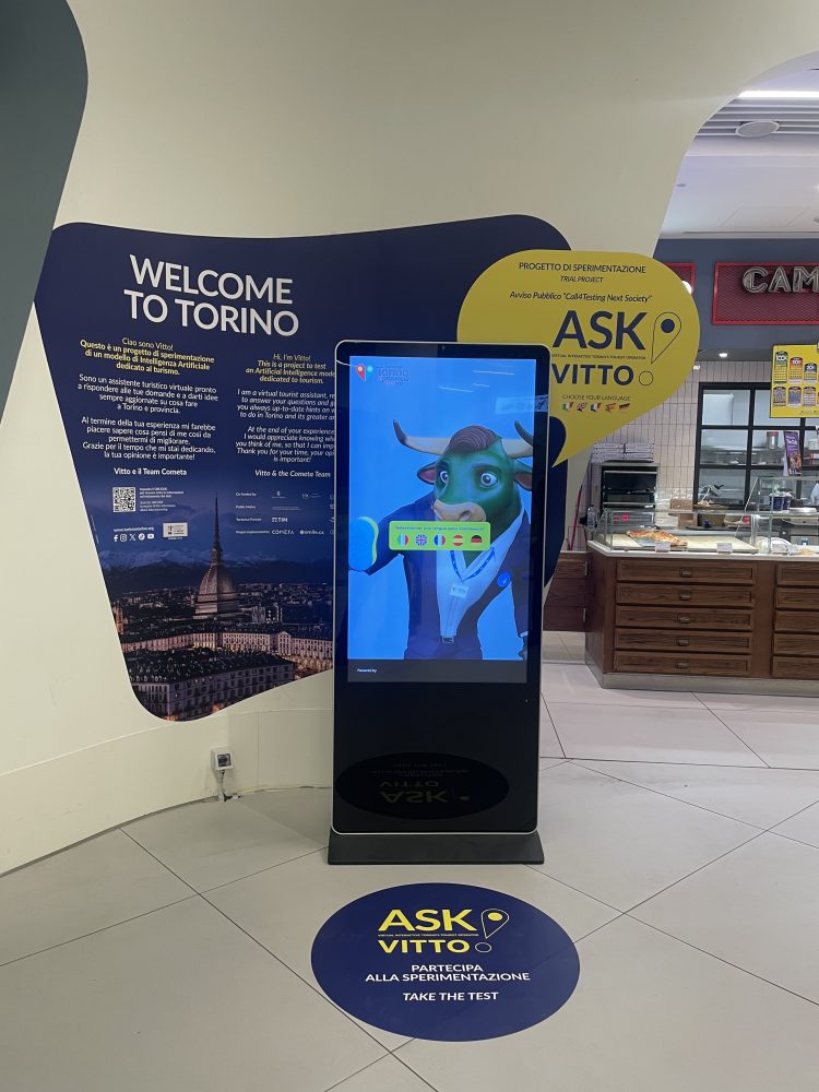 A digital kiosk in a welcoming space displaying "Ask Vitto," an interactive virtual assistant for tourists in Turin.