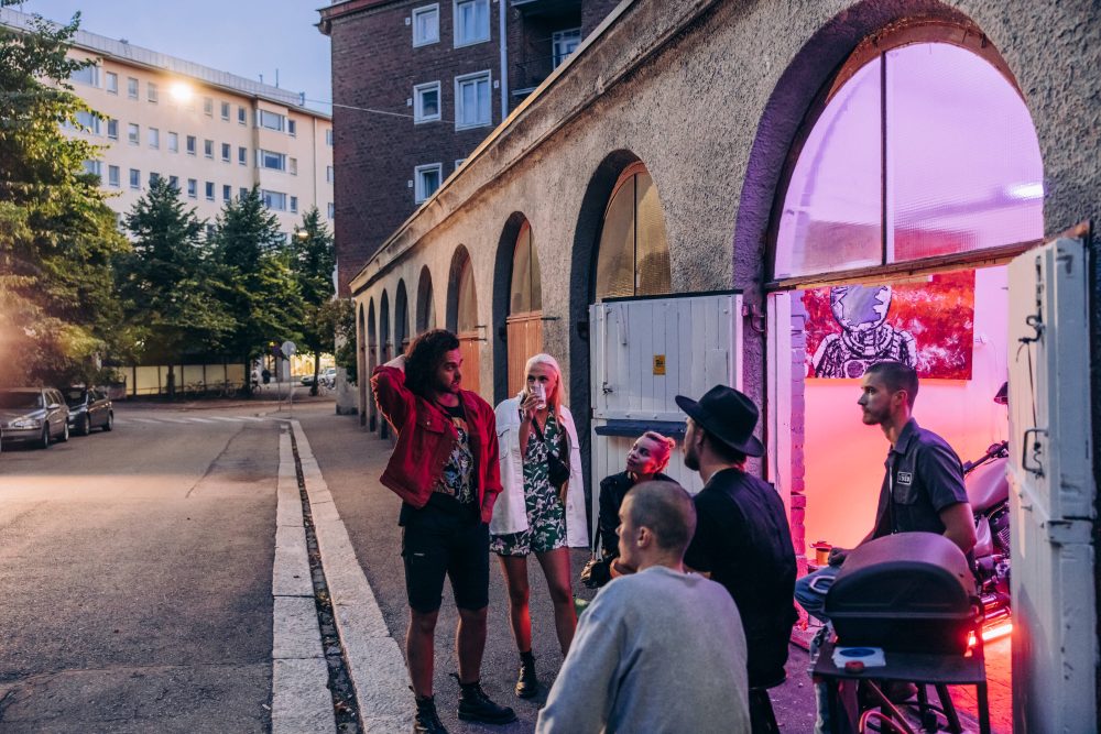 A group of stylish individuals converse outside a venue with colorful lighting, set against an urban backdrop.