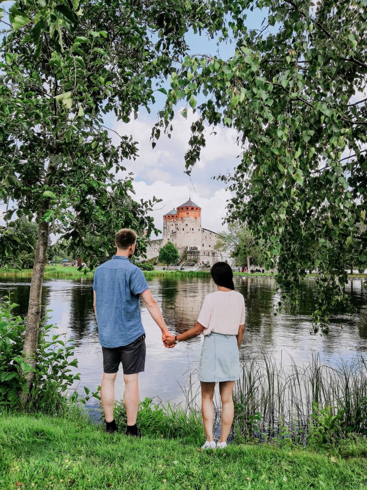 Couple holding hands by the lake, looking at the medieval Olavinlinna Castle in Savonlinna, Finland.