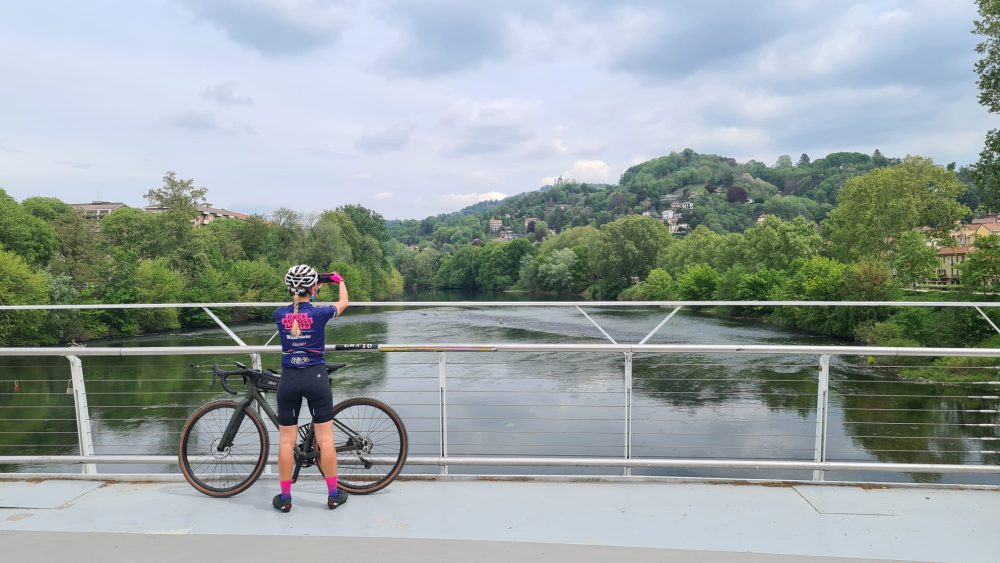 A cyclist in a blue jersey and pink accessories stands on a bridge, capturing the lush, green river landscape with a phone.