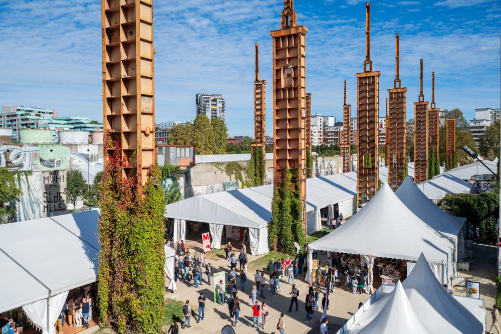 A bustling outdoor event beneath towering rust-colored structures, surrounded by white tents and vibrant graffiti on nearby walls.