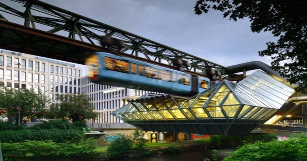 A suspended blue train glides above a modern glass building, set against a backdrop of urban architecture and greenery.