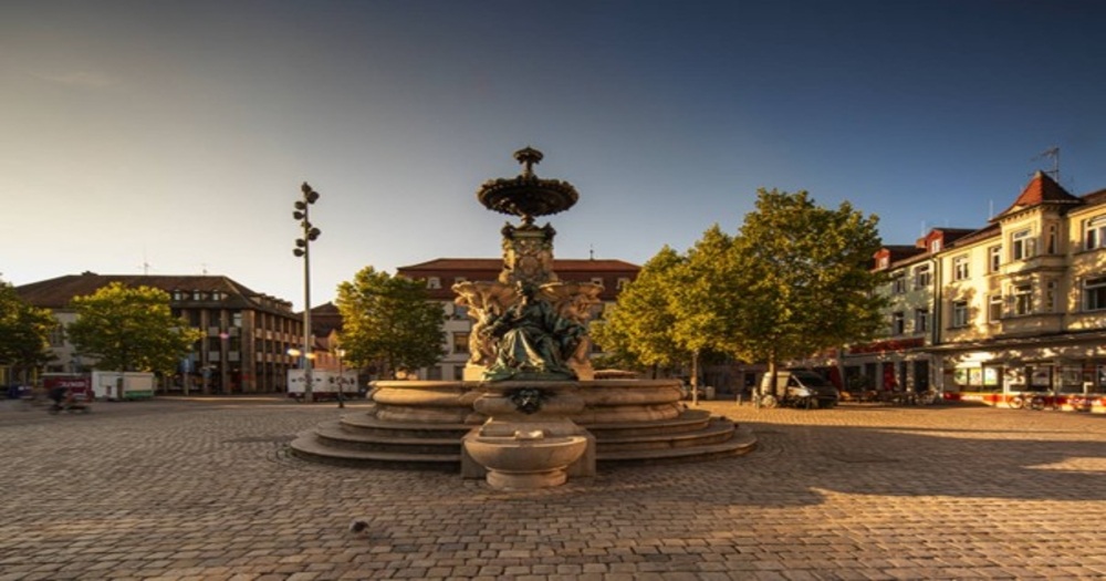 A historic fountain surrounded by trees and buildings, set in a sunny, open square with cobblestone pavement.