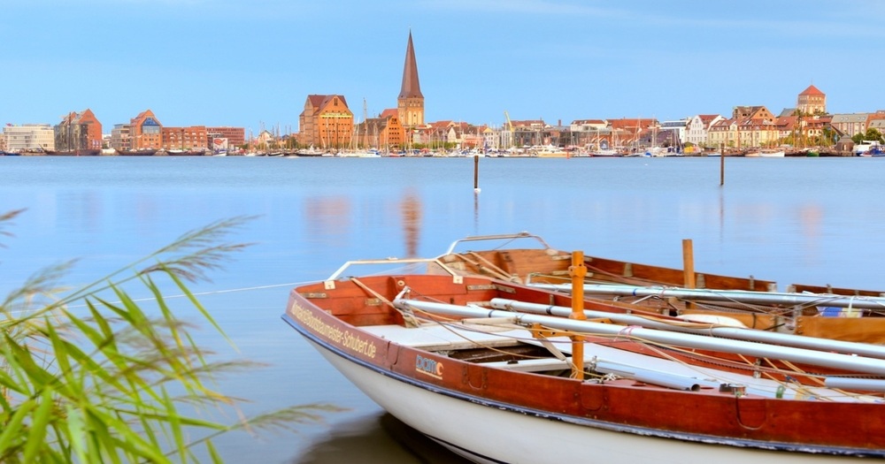 Serene lakeside view with rustic boats in foreground, featuring a skyline of colorful buildings and a church steeple in the background.