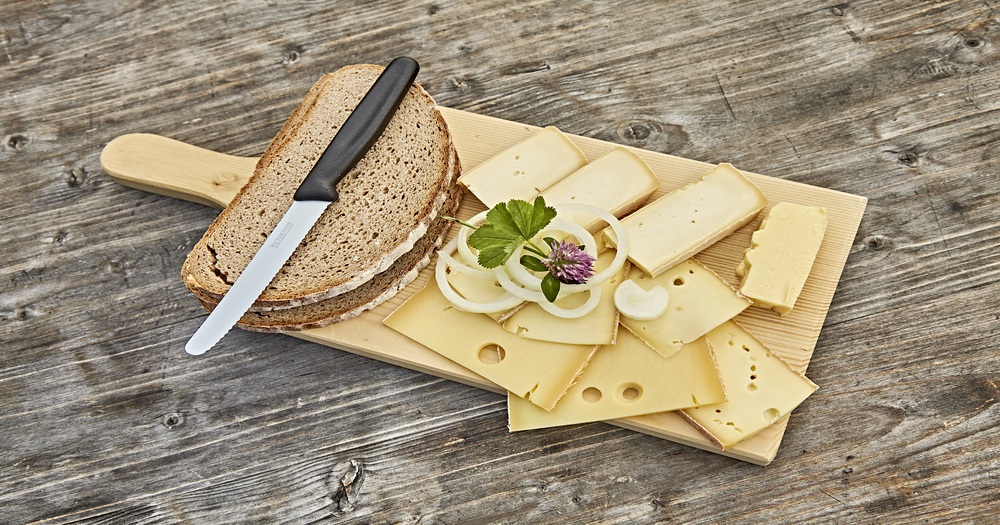 Traditional Alpine cheese platter with rye bread, sliced cheese, onion rings and herbs on a rustic wooden board.