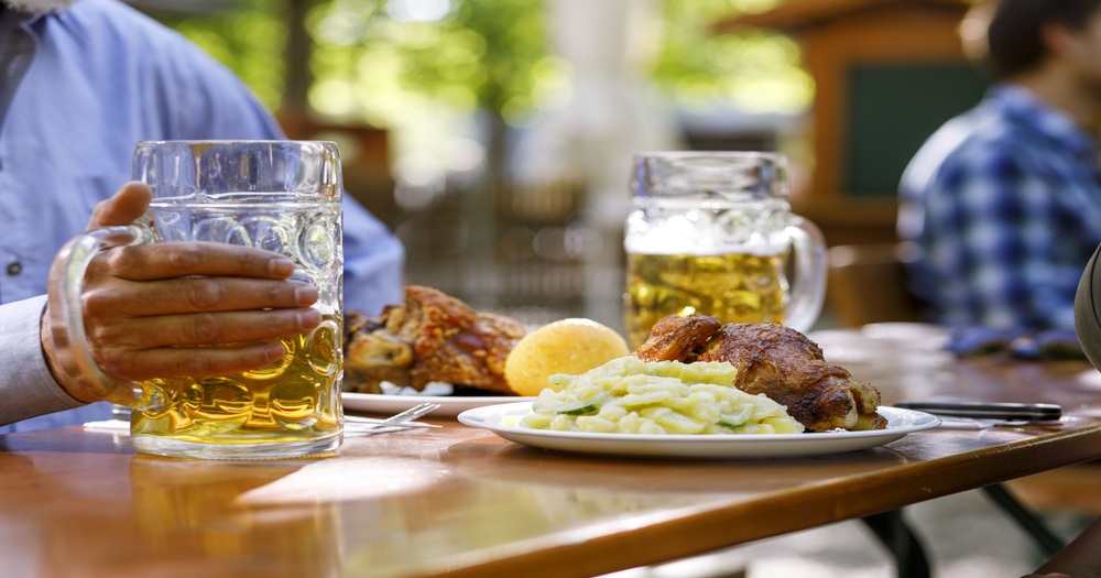 A hand holds a large beer mug beside a plate of roasted chicken, creamy mashed potatoes, and a lemon slice on a sunny outdoor table.