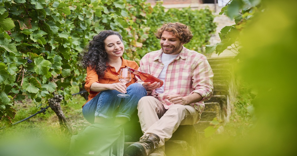A couple enjoys wine together in a vineyard, seated on stone steps surrounded by lush green grapevines.