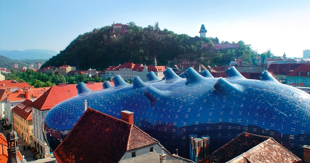A futuristic blue building, the Kunsthaus Graz, contrasts with traditional rooftops and a green hillside in Graz, Austria.