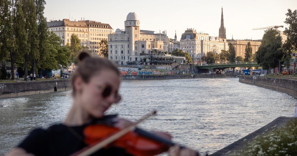 A riverside scene featuring a person playing the violin, with buildings, trees, and a bridge in the background during sunset.