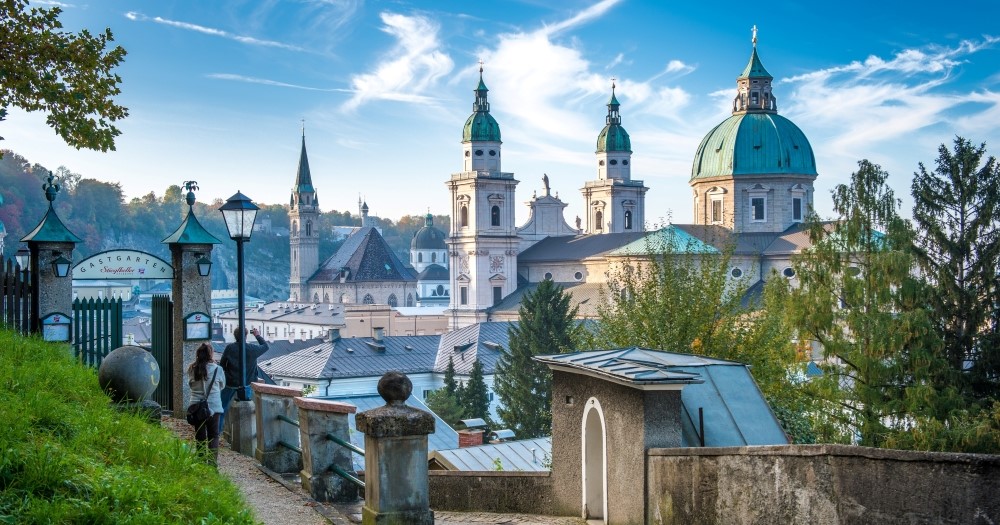 A scenic view of a historic city with green-domed churches and a clear blue sky, framed by trees and street lamps.