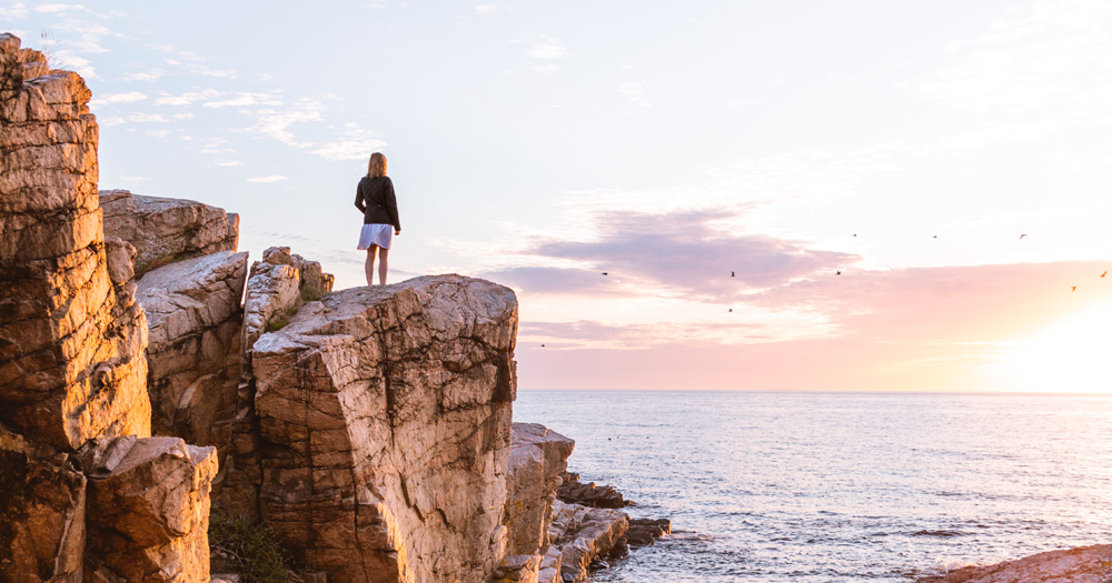Girl standing on coastal rocks at Gudhjem, Bornholm, Denmark, admiring the sea at sunset.
