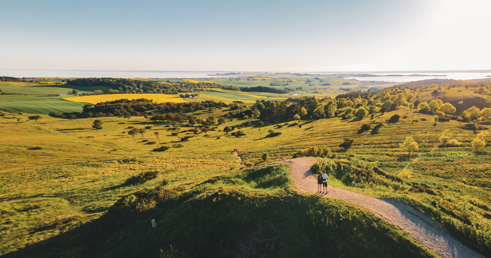 Two hikers stand on a hilltop trail, overlooking a lush green landscape with fields and a serene coastline under a clear sky.