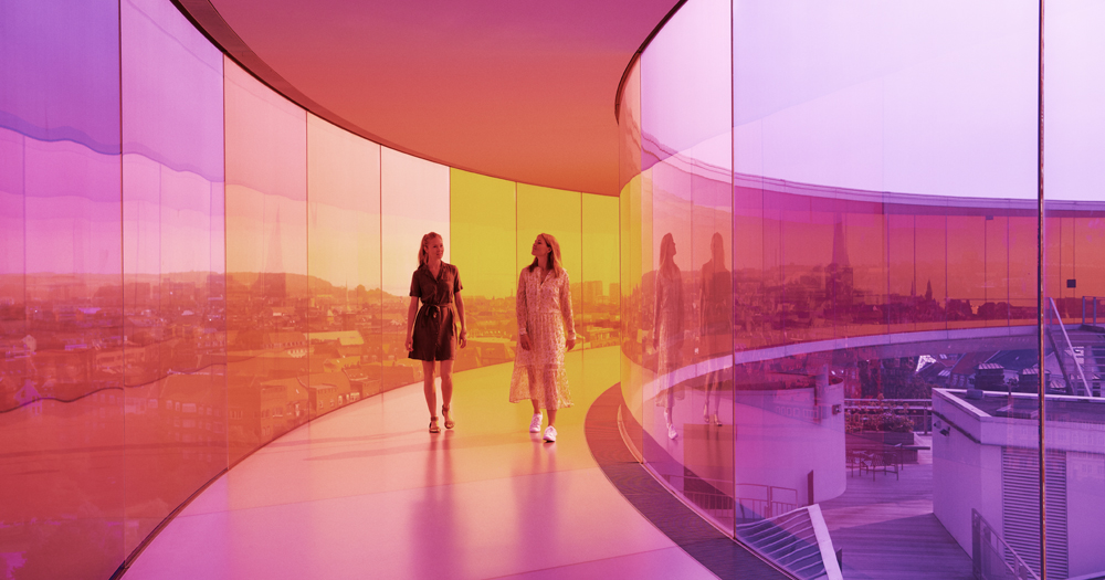 Two women stroll through a brightly colored glass walkway, reflecting a vibrant urban landscape in shades of purple, orange, and yellow.