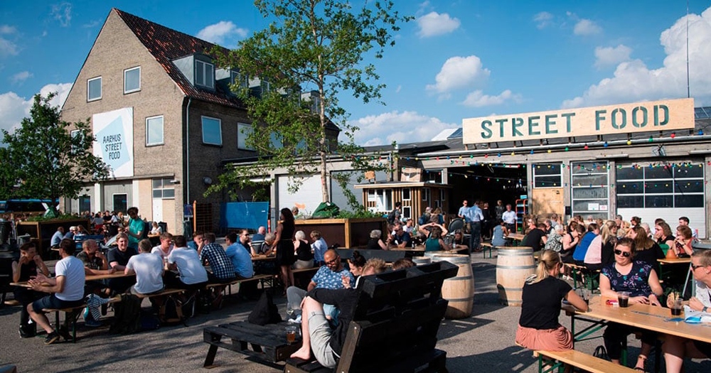 A lively outdoor street food market with people dining at long tables, surrounded by a mix of greenery and a large sign reading "STREET FOOD."