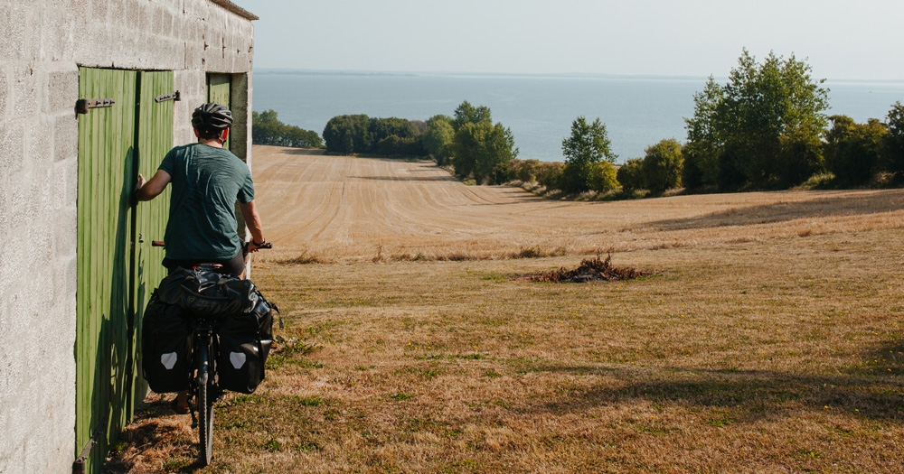Cyclist departing from a green-doored shed, overlooking fields and the sea in Bølgemose, Ærøskøbing, Denmark.