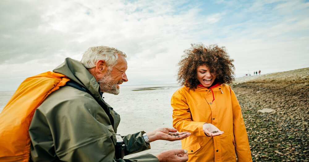 Two people on a rocky beach examine small shells and stones, with cloudy skies and a distant shoreline in the background.