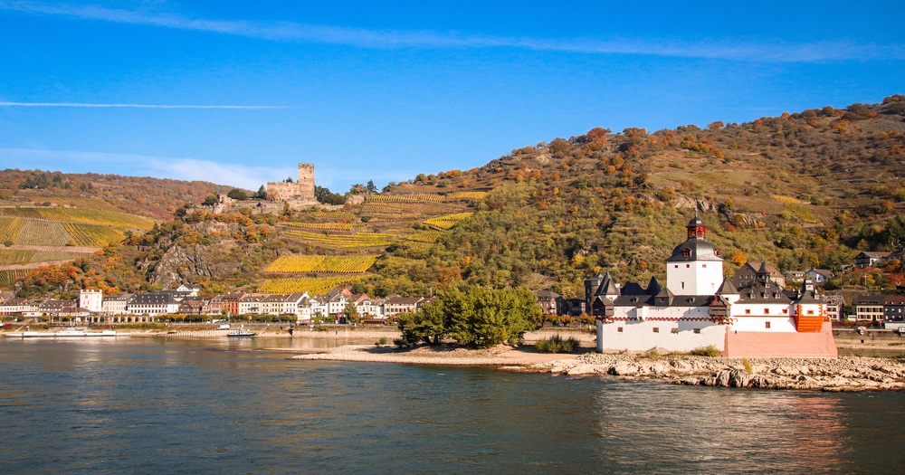 A scenic view of the Rhine River featuring a white castle and a hillside dotted with vineyards, backed by a clear blue sky.