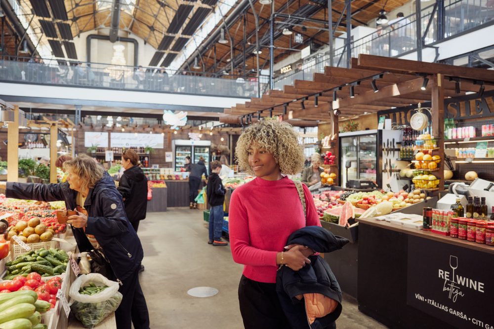 A woman browsing fresh produce stalls inside Riga’s Āgenskalns Market, a lively indoor food market with wooden structures and high ceilings.