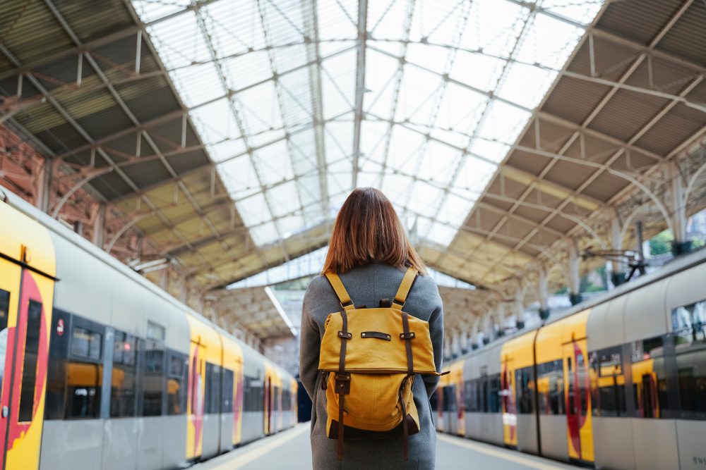 A solo traveler with a mustard yellow backpack stands on a train platform beneath a grand, glass-roofed station, facing two sleek yellow and gray night trains ready for departure.